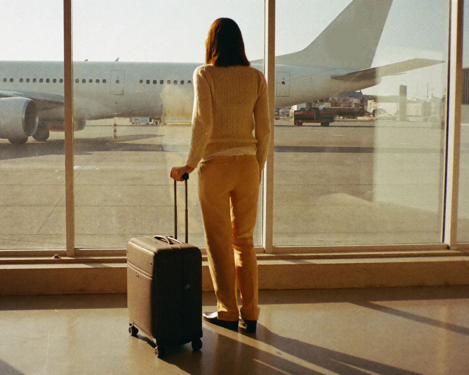 A traveller with a small suitcase at a sunny airport window, looking out at the runway.