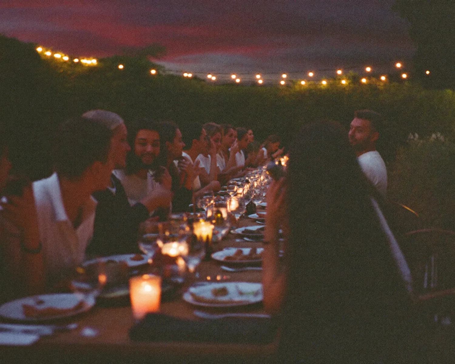 Friends at a long table in a garden at dusk, under string lights, with no phones anywhere.