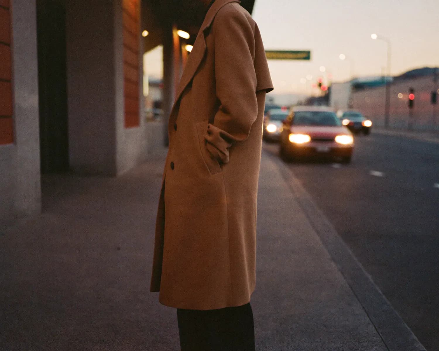 A woman in a camel coat waiting at the kerb at dusk as a car pulls up.