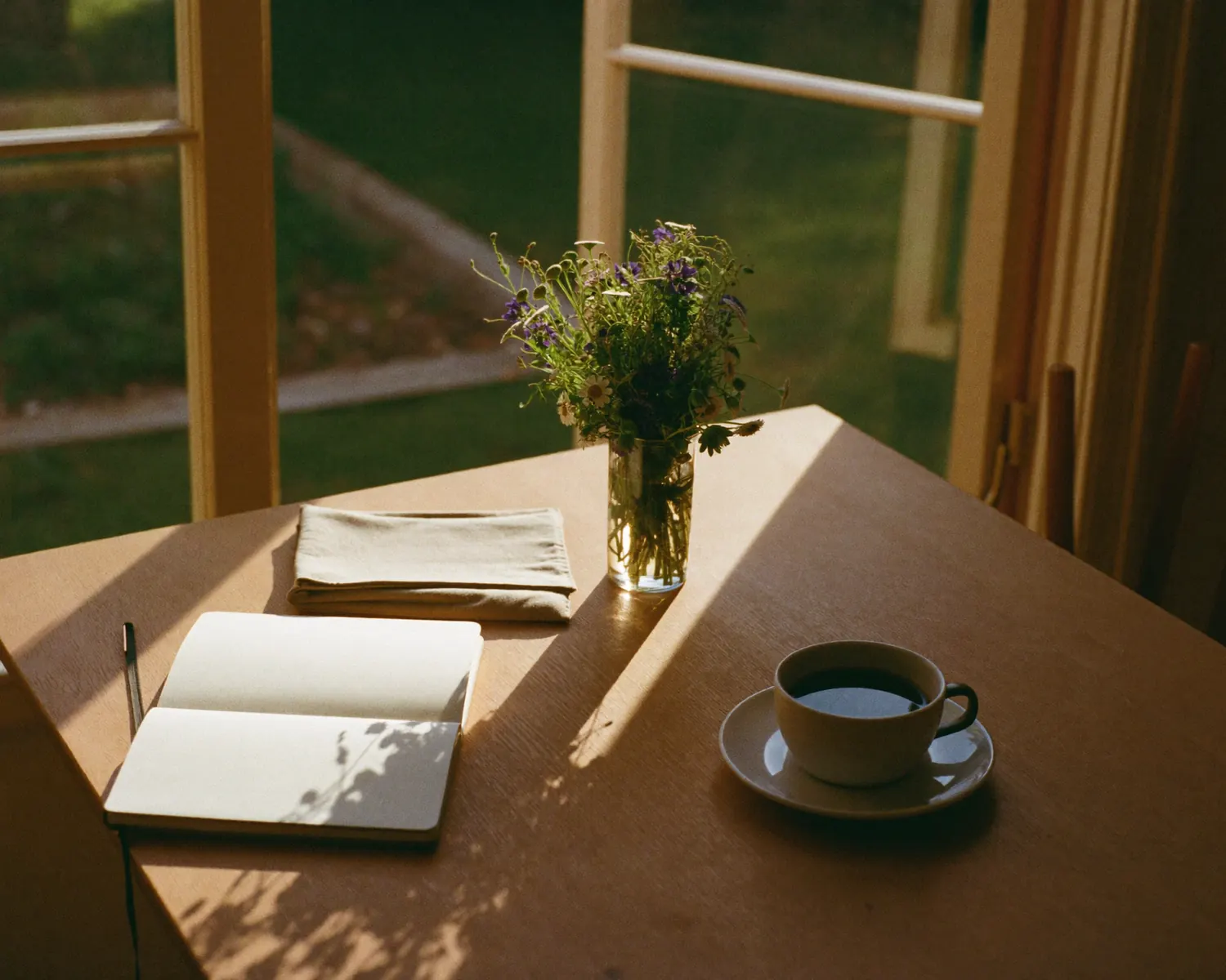 A wooden table by an open window in the afternoon sun, with a notebook, a coffee and wildflowers.
